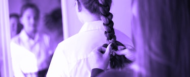 A blurred-out woman braiding a French braid on her daughters head, in front of a mirror.