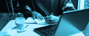 A man sitting at a table with a laptop and writing notes in his notebook