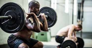 A man in the foreground does a front squat with weights while a man in the background rests.