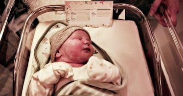 newborn baby in white onesie and grey swaddle in hospital crib