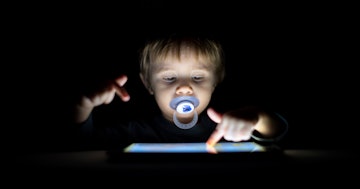 A child with a pacifier in his mouth uses a tablet in the dark.