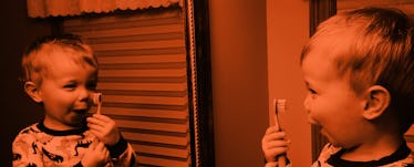 A little kid brushing its teeth while laughing and looking at himself in a mirror