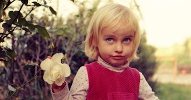 A blonde toddler girl in a white turtleneck and red shirt holding a white rose in a garden