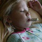 A little blonde girl lying in her PJs in bed and a digital thermometer in her mouth due to coronavir...