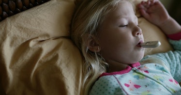 A little blonde girl lying in her PJs in bed and a digital thermometer in her mouth due to Coronavir...
