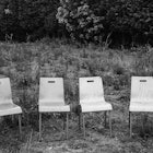 Four wooden chairs stand empty in an overgrown field