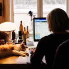 A work from home parent sitting and looking at her computer with a cat lying on the desk 