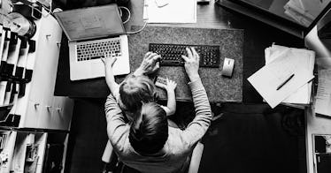 A father sitting at his work desk with his kid in his lap while both of the use keyboards in front o...