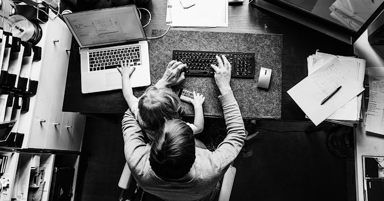 A father sitting at his work desk with his kid in his lap while both of the use keyboards in front o...