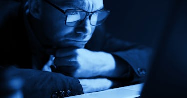 Man wearing blue light blocking glasses with head resting on fists frowning at computer screen cover...
