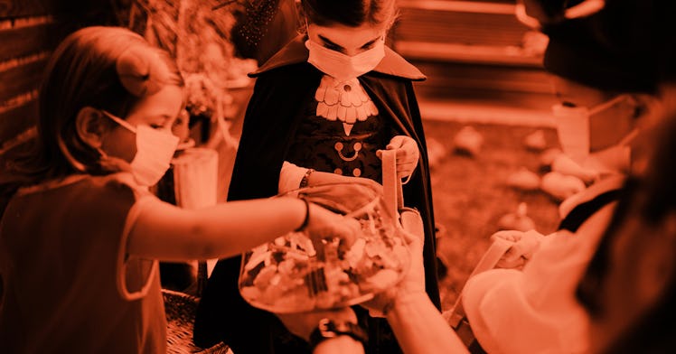 A group of three children taking candy out of a bucket for Halloween, wearing costumes and Covid-19 ...