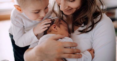 a baby cradled in a woman's arms, with a toddler placing her hand on the baby's head, depicting baby...