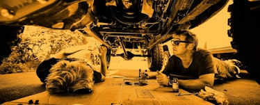 A dad and his son under a car looking at the different parts of the car and fixing it 