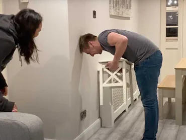 A man attempts the chair challenge at home while his wife looks on.