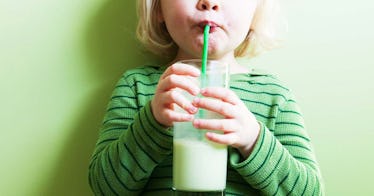 A child in a green shirt drinking a greenish protein shake for kids through a straw, leaning against...