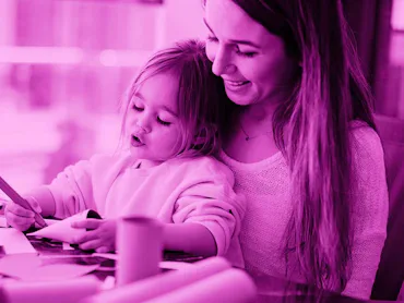 pink photo edit of a young girl working on construction paper crafts while sitting in her mother's l...
