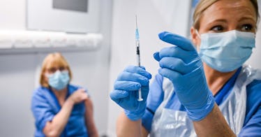 Two women in scrubs are masked; one holds a vaccine in her gloved hands