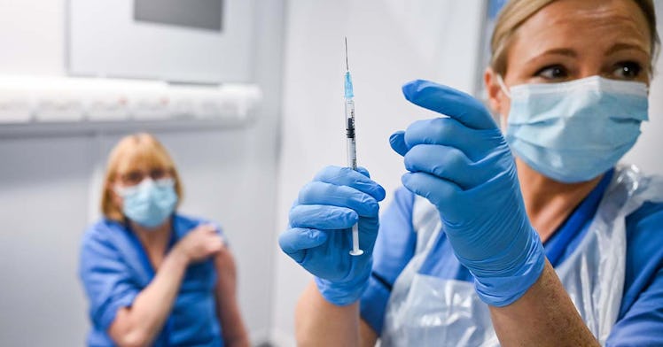 Two women in scrubs are masked; one holds a vaccine in her gloved hands