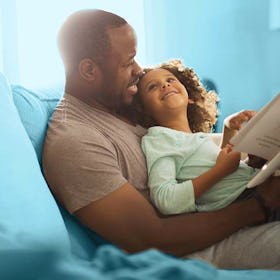 A father sitting on a blue couch with his daughter in his lap, reading her a book of poems