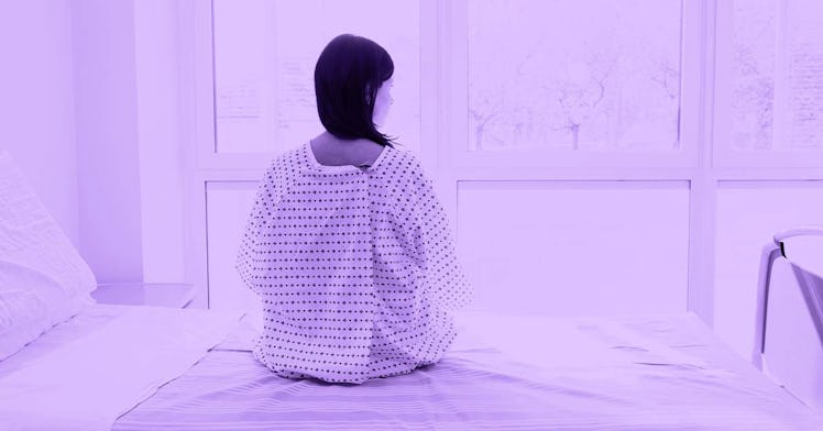 A black haired woman in a hospital gown sits on a bed and looks out a window.