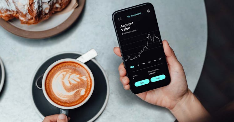A hand holding a phone checking bitcoin prices next to a latte and a croissant