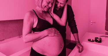 An older woman with smile lines looking at her pregnant belly with an older man in a bathroom settin...
