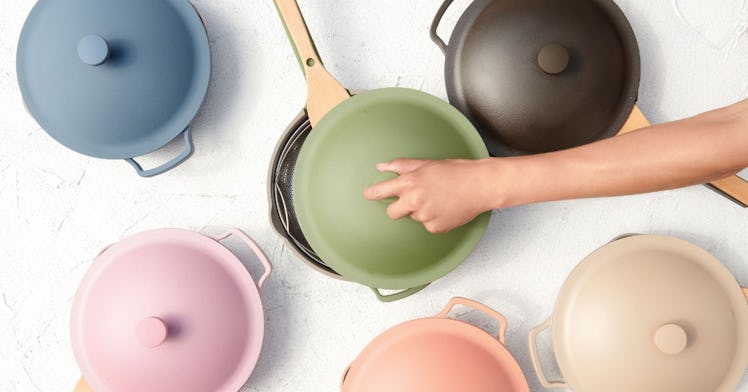 A hand opens the lid of an Always Pan by Our Place among multiple colors of the pan