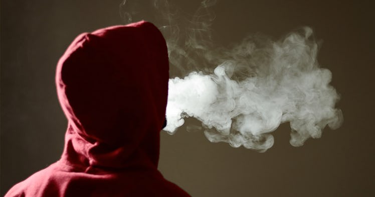 A teen in a hoodie facing away, blowing marijuana smoke.