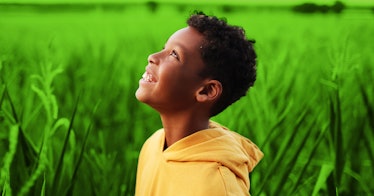 A black child in a yellow hoodie smiling as he stands in a field looking at the sky.