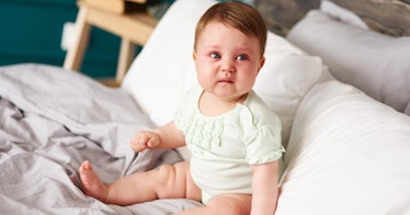 An infant baby sits on a bed, in tears