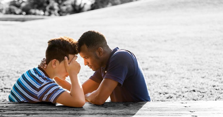 A father and son sit together at a picnic table with foreheads touching while the son covers his eye...