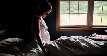 A pregnant person sitting on a bed in the dark.