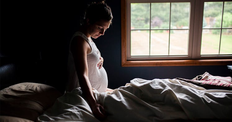 A pregnant person sitting on a bed in the dark.