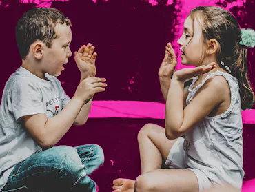 Young boy and girl playing Pat-a-Cake in front of a pink background.