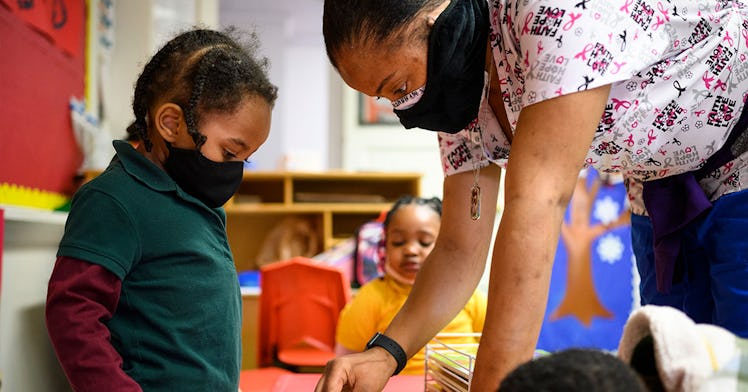 A day care worker stands, bent over, a child. Both wear masks