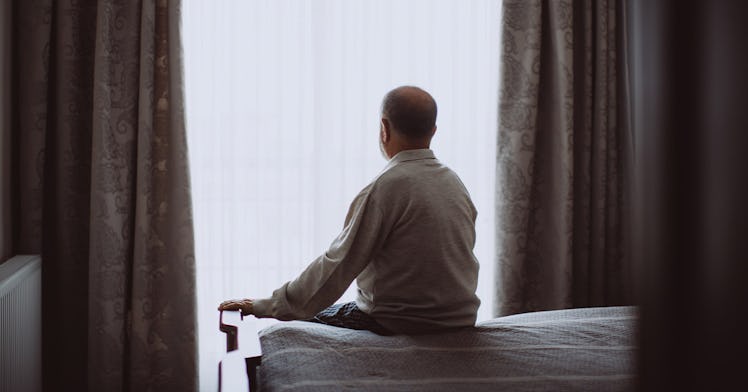 An old man sits on a bed and looks out a covered window