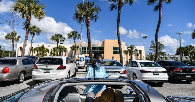 A person in a mask gives a COVID test in a parking lot. Palm trees are in the background.