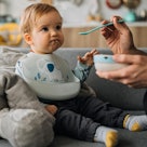 A parent feeds a baby a spoonful of Cheerios from a bowl.