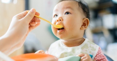 an Asian baby gets fed food from a yellow spoon and gazes at parent, off-screen