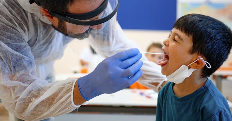 A nurse in full PPE administers a throat swab COVID test to a kid in a classroom