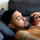 A shirtless new dad holds his naked baby to his chest as they ly on a couch.