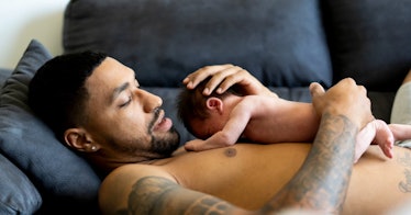 A shirtless new dad holds his naked baby to his chest as they ly on a couch.