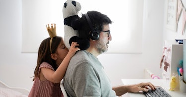 a dad works on his computer remotely while his daughter plays behind him
