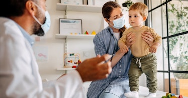 A mom wearing a mask holds her toddler on an exam table as a doctor approaches.
