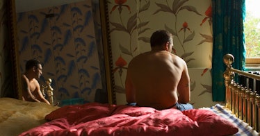 A shirtless man sits on a bed, his image reflected in a mirror.