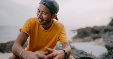 A man in a backwards hat sits outdoors in a rocky landscape and smiles.
