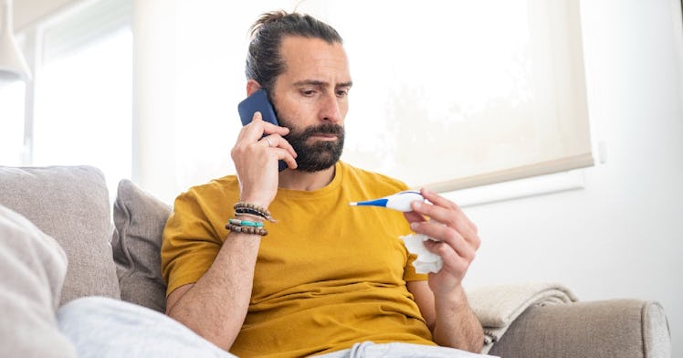A man sits on a couch and holds a thermometer and a tissue.