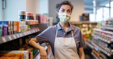 a woman stands in a store she works at with a mask on