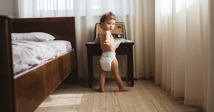 A 15-month-old in a diaper stands in their parent's bedroom.
