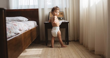 A 15-month-old in a diaper stands in their parent's bedroom.
